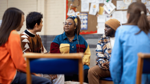 A diverse group of six people sitting in a circle, engaged in conversation. They are in a room with a bulletin board in the background. The setting suggests a casual or support group meeting.