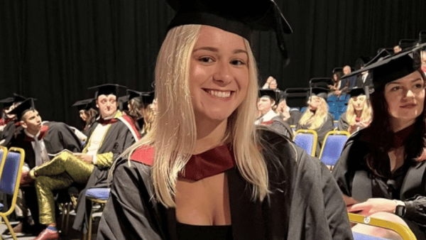 A graduate smiles while wearing a cap and gown, sitting in an auditorium with other graduates in the background.