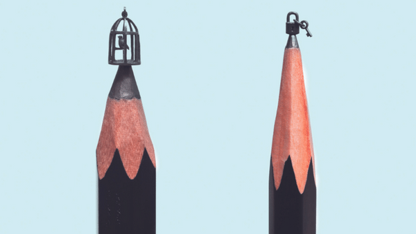 Two sharpened pencil tips with intricate sculptures: one depicts a small birdcage with a tiny bird, and the other shows a padlock. Both are carefully carved from the graphite.