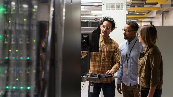 Three people in a server room examine a computer monitor. One person gestures towards the screen while the others attentively watch. Nearby, server racks with illuminated lights are visible. All are casually dressed.