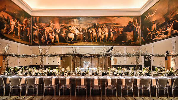 An opulent banquet hall features a long dining table adorned with elegant flower arrangements and numerous candles. The walls are decorated with large, classical paintings. Transparent chairs line the table, and soft lighting creates a warm atmosphere.