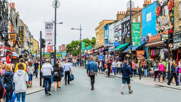 A busy street scene with numerous people walking on a vibrant road lined with colorful shops and storefronts. Signs advertising deals and various store names are visible. Buildings display unique designs and the atmosphere is lively and bustling.