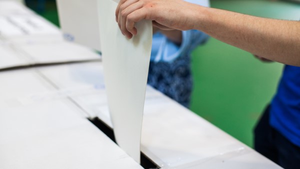 A person is placing a white ballot paper into a slot on a white ballot box. Another person stands nearby. The background is green and blurred.