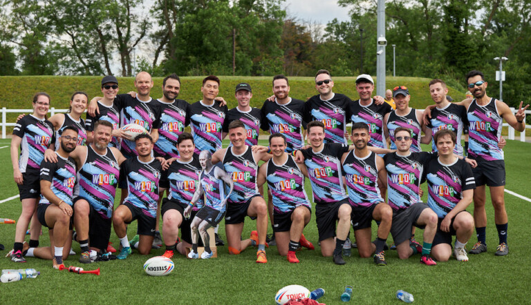 A group of 23 people, wearing colorful sports jerseys with "WOLOP" on them, pose for a photo on a grassy sports field. Some kneel in the front row while others stand behind them. Rugby balls are visible on the ground. Trees are in the background.