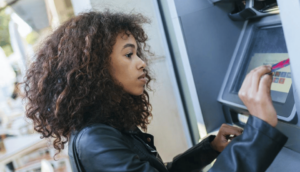 A person with curly hair and a black jacket uses a touchscreen ATM, holding a red card. They appear focused while interacting with the machine.