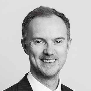 Black and white portrait of a smiling man in a suit and tie. He has short hair and is looking directly at the camera against a plain background.