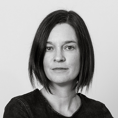 Black and white portrait of a person with shoulder-length hair, wearing a dark top, against a plain background. The person has a neutral expression and is looking directly at the camera.