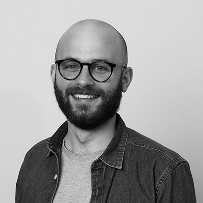 A man with a beard and glasses smiles at the camera. He is wearing a denim shirt over a light t-shirt. The image is in black and white.