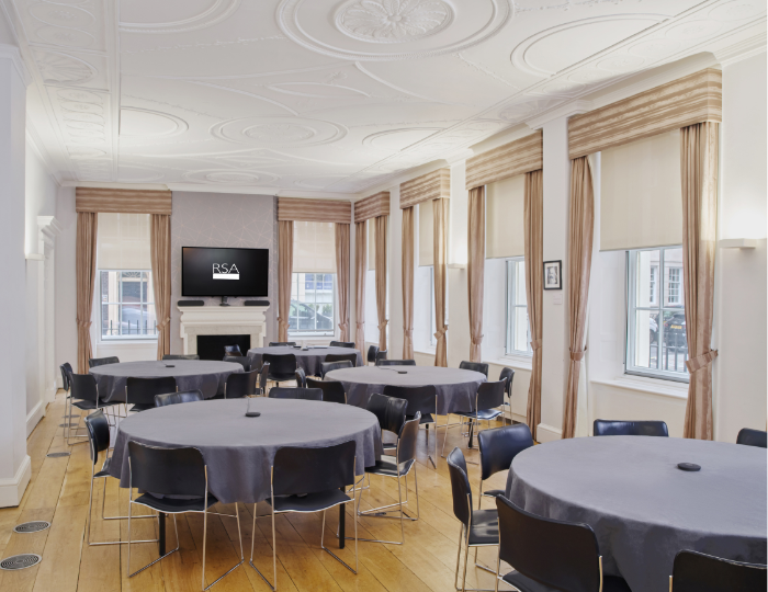 A bright conference room with round tables covered in gray tablecloths and surrounded by black chairs. Large windows with beige curtains line the wall. A screen displaying "RSA" is mounted above a fireplace. The ceiling features ornate designs.