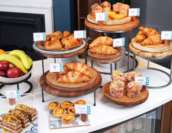 A display of various pastries, including croissants, danishes, and custard tarts, on wooden and metal stands. Fresh fruit like bananas and apples are on the side. Each pastry has a small label indicating its name.