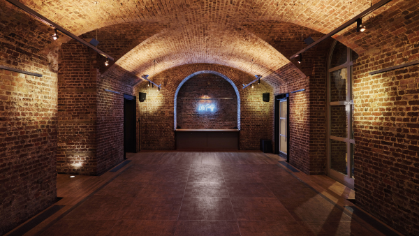 A dimly lit room with arched brick ceilings and walls. The space features recessed lighting and a neon sign at the far end. The floor is tiled, and there are subtle reflections on the surface. No furniture visible.