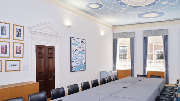 A conference room with a long table surrounded by black chairs. The ceiling has intricate artwork with a light blue background. Walls are adorned with framed pictures and a large window lets in natural light. A wooden door is on the left.
