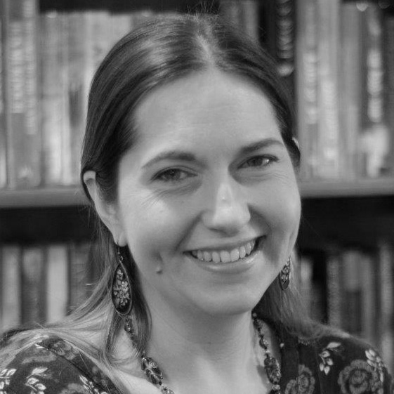 A black and white portrait of a smiling person with long hair, wearing earrings and a patterned top. They are in front of a blurred bookshelf background.