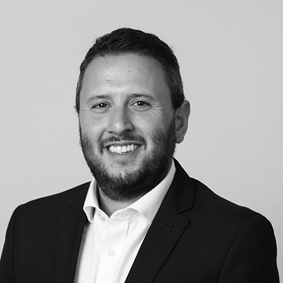 A black and white portrait of Tom Stratton with short hair and a beard, smiling. He is wearing a white shirt and a dark suit jacket. The background is plain.