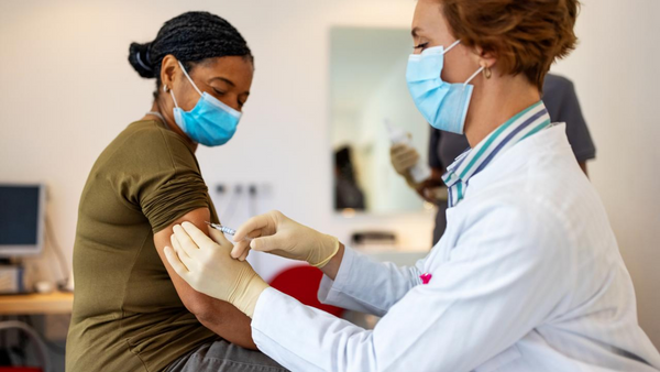 A healthcare professional in a white coat and gloves administers a vaccine to a seated woman. Both are wearing face masks. A third person is blurred in the background. A desk with a computer is visible in the room.