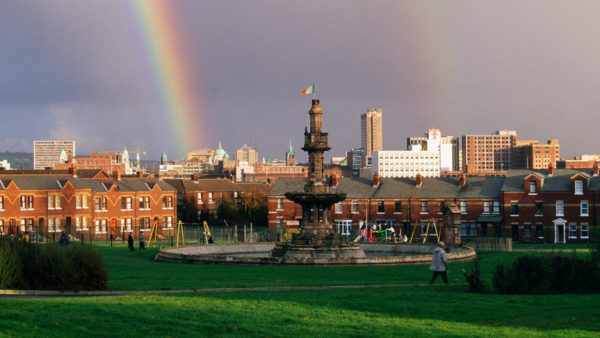 A park with a historic fountain and lush green grass. In the background, a row of red-brick houses and a city skyline with various buildings under a cloudy sky. A vibrant rainbow arcs across the sky. People walk and children play nearby.