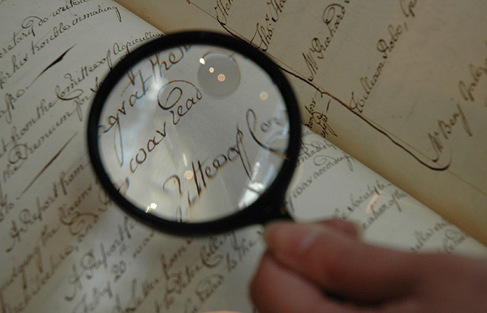 A hand holding a magnifying glass over a handwritten document. The magnified text shows elegant cursive writing on aged paper, enhancing the clarity of the script.