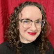 A person with curly hair and glasses smiles while wearing a black top. The background is a red curtain.