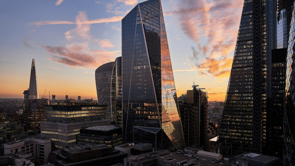 A cityscape at sunset showcases modern skyscrapers with glass facades reflecting the colorful sky. Tall, angular buildings dominate the skyline, and wispy clouds are illuminated by the orange and pink hues of the setting sun.