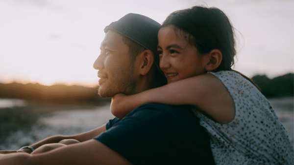 A man wearing a cap is sitting with a young girl who is smiling and wrapping her arms around his shoulders. They are outdoors during sunset, and the background shows a blurred landscape with trees and sky.