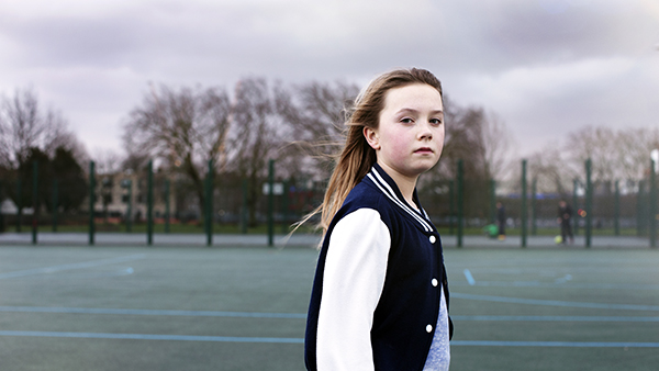 A young girl with long hair stands on an outdoor sports court, wearing a black and white varsity jacket. The background features trees and a cloudy sky.