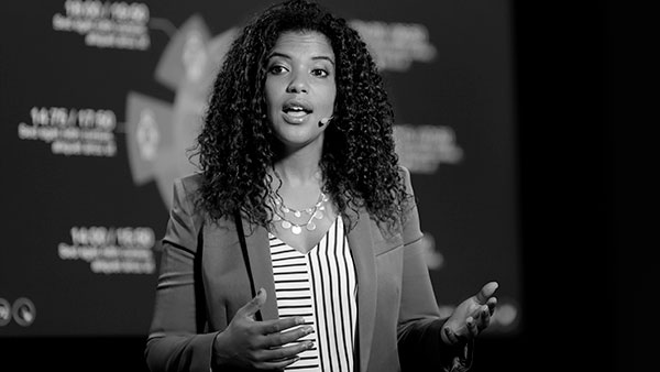 A woman with curly hair speaks on stage, wearing a blazer over a striped shirt. She gestures with her hand, and a microphone is visible on her cheek. The background displays blurred text and symbols. The image is in black and white.