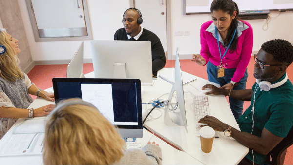 Five people are working in an office, each at a computer. Four are seated around a table with screens, while one person stands and talks. Headphones, books, and a coffee cup are visible on the table. The room has white walls and red carpet.