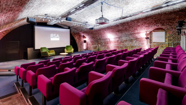 An auditorium with rows of purple seats facing a screen displaying "RSA." The room has arched brick walls and ceiling, with two green chairs and a small round table on stage. Ceiling lights are turned on.