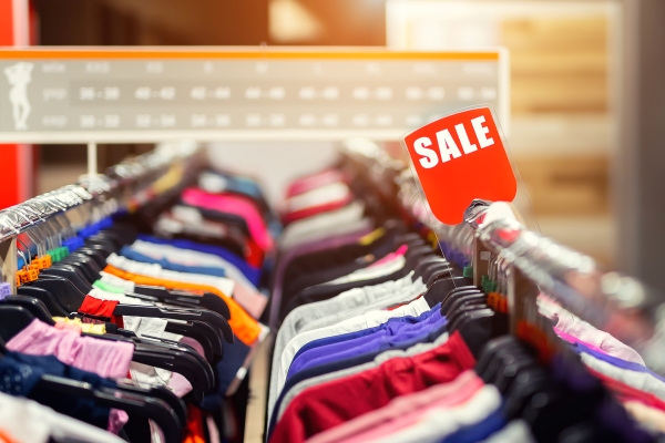A clothing rack filled with various colorful garments on hangers, with a red "SALE" sign prominently displayed. The scene is in a store with soft lighting, suggesting a discount or promotion event.