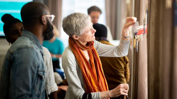 A group of people are gathered indoors. An older woman with short gray hair and an orange scarf is pinning a card to a string with clothespins. Others, including a man with a beard and glasses, watch attentively.