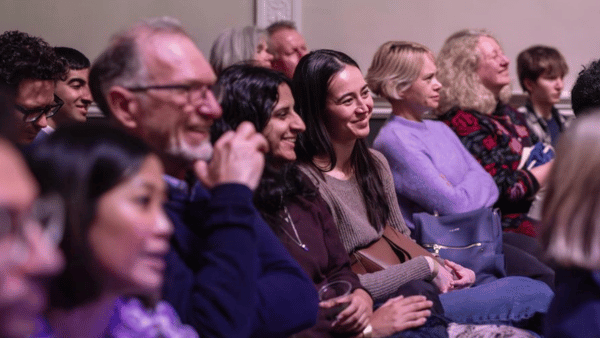 A diverse group of people sitting in an audience, smiling and listening attentively. The setting appears to be a seminar or presentation. Everyone is dressed casually, and the atmosphere seems engaging and positive.