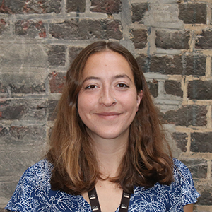 A person with shoulder-length brown hair and a nose ring smiles in front of a textured brick wall. They wear a blue patterned top.