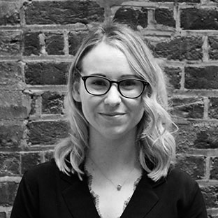 A smiling person with shoulder-length hair and glasses is wearing a dark top. They are standing in front of a brick wall. The image is in black and white.