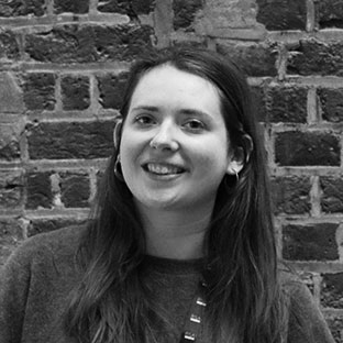 Smiling person with long hair, wearing a dark sweater, standing in front of a brick wall. The image is in black and white.