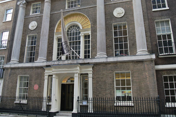A historic brick building with tall columns and arched windows. It features decorative medallions on the facade and a central entrance with steps leading up to a doorway. A flag hangs above the entrance, and a black fence lines the front.