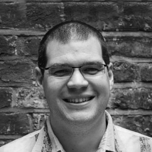 A black and white photo of a person with short dark hair and glasses, smiling. They are wearing a patterned shirt and standing in front of a textured brick wall.