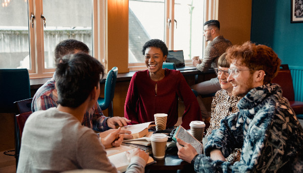 A group of people sit around a table, engaged in conversation and smiling. They have notebooks and coffee cups in front of them. The background shows large windows and soft lighting, creating a cozy atmosphere.