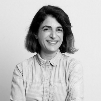 A person with shoulder-length hair smiles at the camera. They are wearing a long-sleeved, button-up shirt with vertical stripes. The background is plain white.