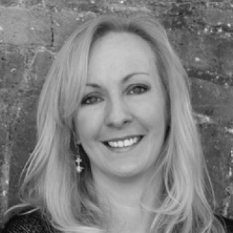 Smiling woman with long blonde hair, wearing a cross-shaped earring, against a brick wall background. Black and white image.