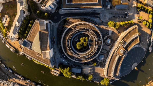 Aerial view of a cityscape featuring a circular building complex surrounded by roads and greenery. Several boats are visible on the adjacent river, and other modern structures are nearby. The scene is bathed in warm sunlight.