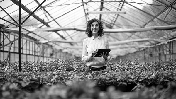A person with curly hair, wearing a sweater, stands smiling with a tablet in their hands. They are surrounded by potted plants inside a large greenhouse with a glass roof. The image is in black and white.