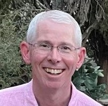 Smiling man with short white hair and glasses, wearing a pink shirt, stands outside in front of greenery.