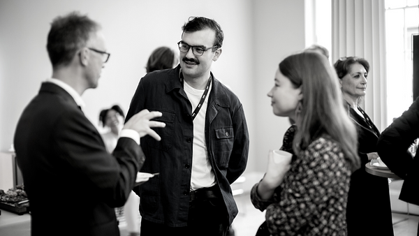 A black-and-white photo shows a group of people in conversation at an indoor event. A man in glasses and a jacket is speaking to a woman holding a cup. Others are engaged in dialogue in the background.