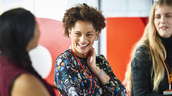 Three people are engaged in conversation. The central person is smiling and wearing a floral-patterned dress with a lanyard. The background is slightly blurred with red and white colors.
