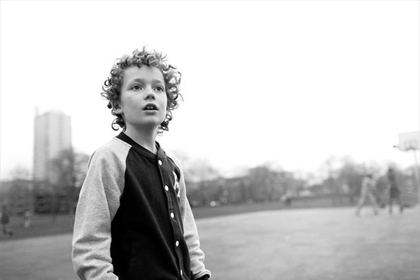 Black and white photo of a curly-haired child wearing a baseball-style shirt, looking upward with an open mouth. A tall building and a basketball hoop are in the background in a park setting.