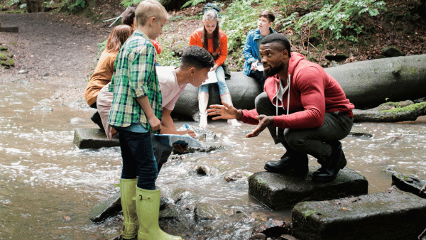A group of children and a man are outdoors by a creek. The man is crouching on a rock, speaking to a boy in a plaid shirt and green boots. Other children are sitting on rocks and logs, observing. The setting is a wooded area.