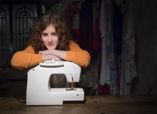 A person with curly hair, wearing an orange sweater, leans over a sewing machine on a wooden table. The background is dimly lit, showing clothing hanging on racks.