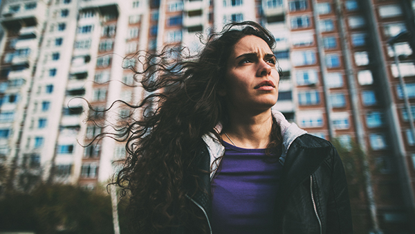 A woman with long, curly hair stands outdoors in front of a tall apartment building. She looks to the side with a thoughtful expression. She is wearing a black jacket over a purple shirt. The sky is overcast.