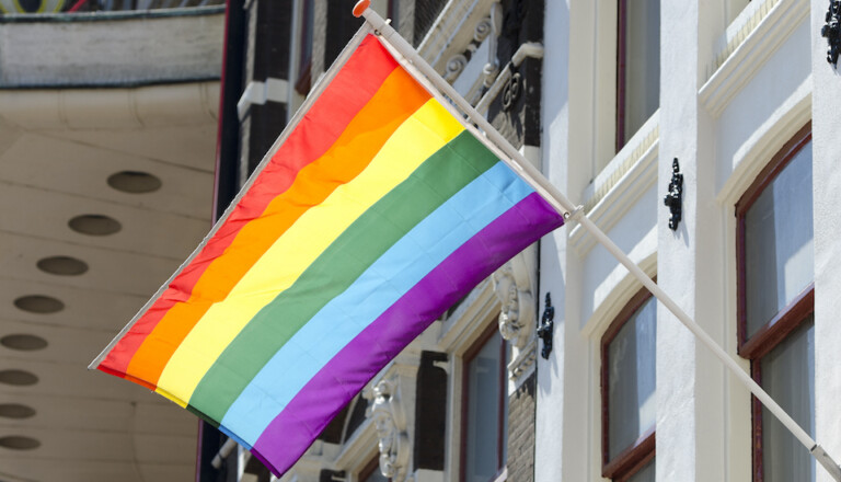 A rainbow flag with red, orange, yellow, green, blue, and purple stripes is flying outside a building with white and beige walls and several windows.