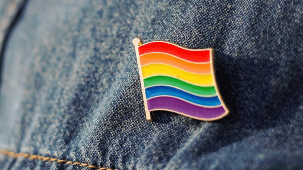 A small pin shaped like a waving rainbow flag is attached to blue denim fabric. The flag features horizontal stripes in red, orange, yellow, green, blue, and purple, symbolizing LGBTQ+ pride.